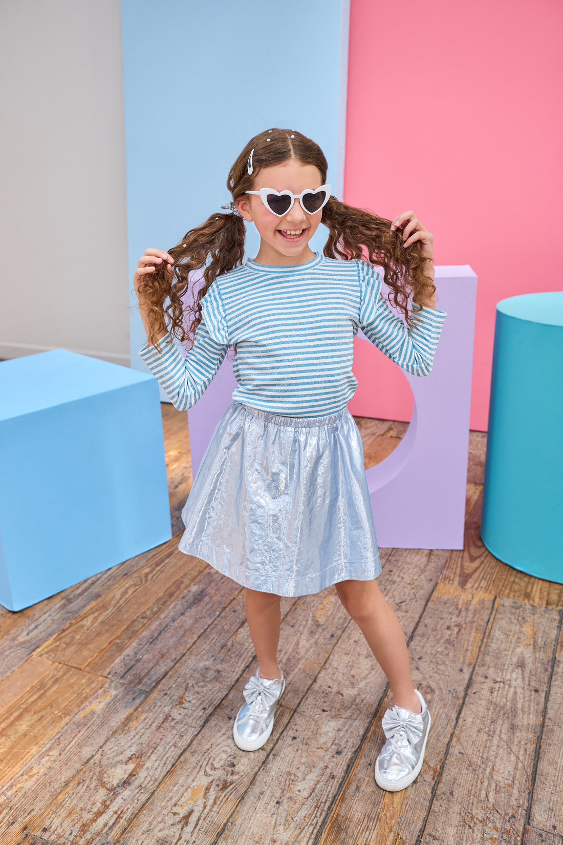 Young girl in a striped shirt and metallic skirt standing in front of colorful geometric shapes on a wooden floor.