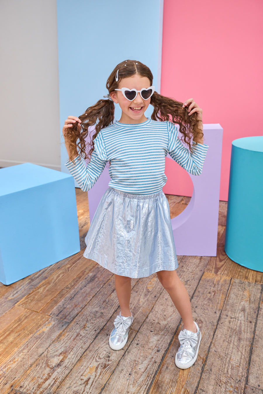 Young girl in a striped shirt and metallic skirt standing in front of colorful geometric shapes on a wooden floor.