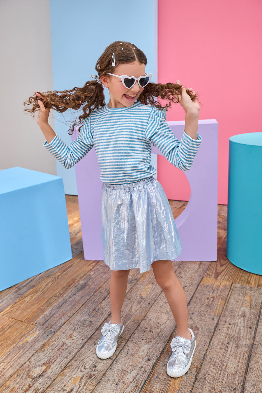 Young girl playing with her hair against a colorful geometric background wearing a blue metallic skirt and blue and white striped long sleeve top.