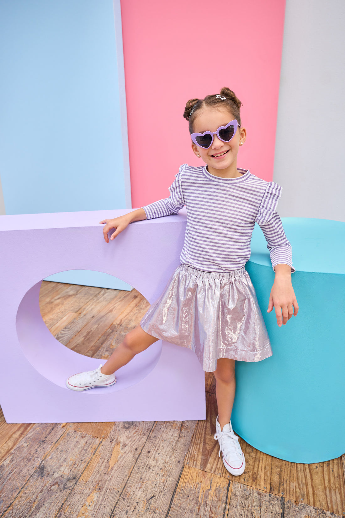Child in a striped shirt and metallic skirt standing against colorful geometric shapes.