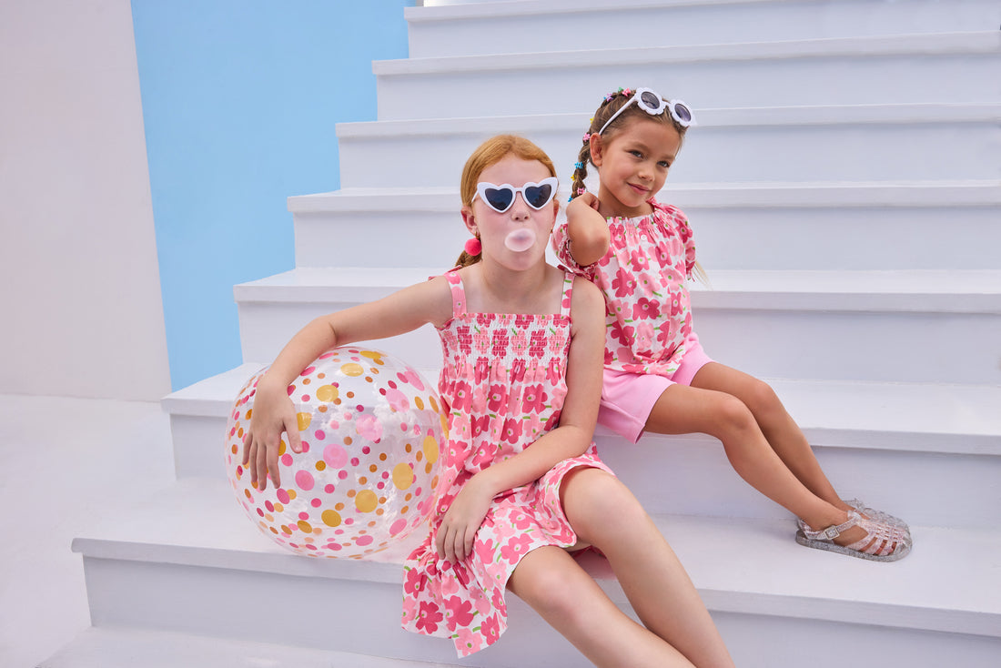 Model on right can be seen wearing a woven top that features elastic neckline, button closure down front, and faux ties on elastic sleeve cuffs for an elevated look. The big red/pink floral print matches back perfectly with the bubblegum knit shorts to complete the look. Model on left can be seen wearing the same floral print but in a woven dress that features faux bow ties on straps and smocking across bust. 