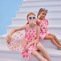 Model on right can be seen wearing a woven top that features elastic neckline, button closure down front, and faux ties on elastic sleeve cuffs for an elevated look. The big red/pink floral print matches back perfectly with the bubblegum knit shorts to complete the look. Model on left can be seen wearing the same floral print but in a woven dress that features faux bow ties on straps and smocking across bust. 