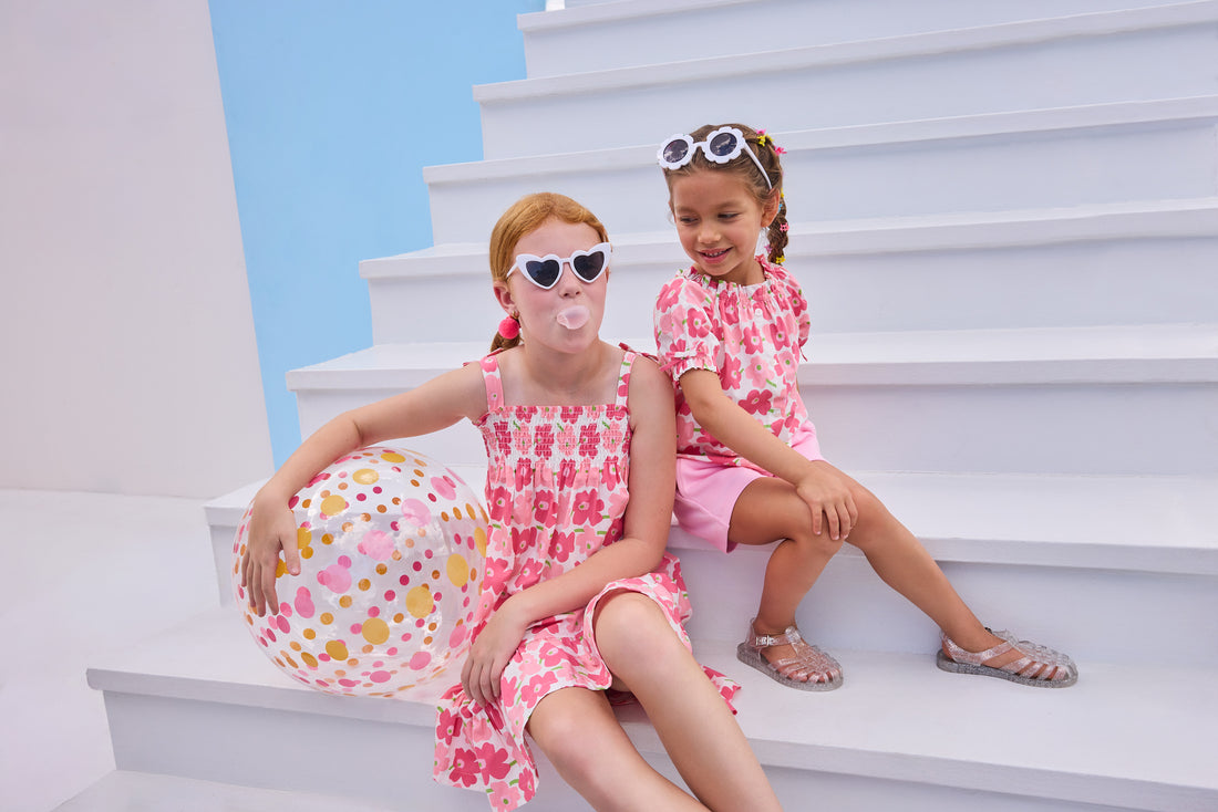 Model on right can be seen wearing a woven top that features elastic neckline, button closure down front, and faux ties on elastic sleeve cuffs for an elevated look. The big red/pink floral print matches back perfectly with the bubblegum knit shorts to complete the look. Model on left can be seen wearing the same floral print but in a woven dress that features faux bow ties on straps and smocking across bust. 