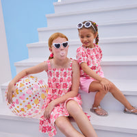 Model on right can be seen wearing a woven top that features elastic neckline, button closure down front, and faux ties on elastic sleeve cuffs for an elevated look. The big red/pink floral print matches back perfectly with the bubblegum knit shorts to complete the look. Model on left can be seen wearing the same floral print but in a woven dress that features faux bow ties on straps and smocking across bust. 