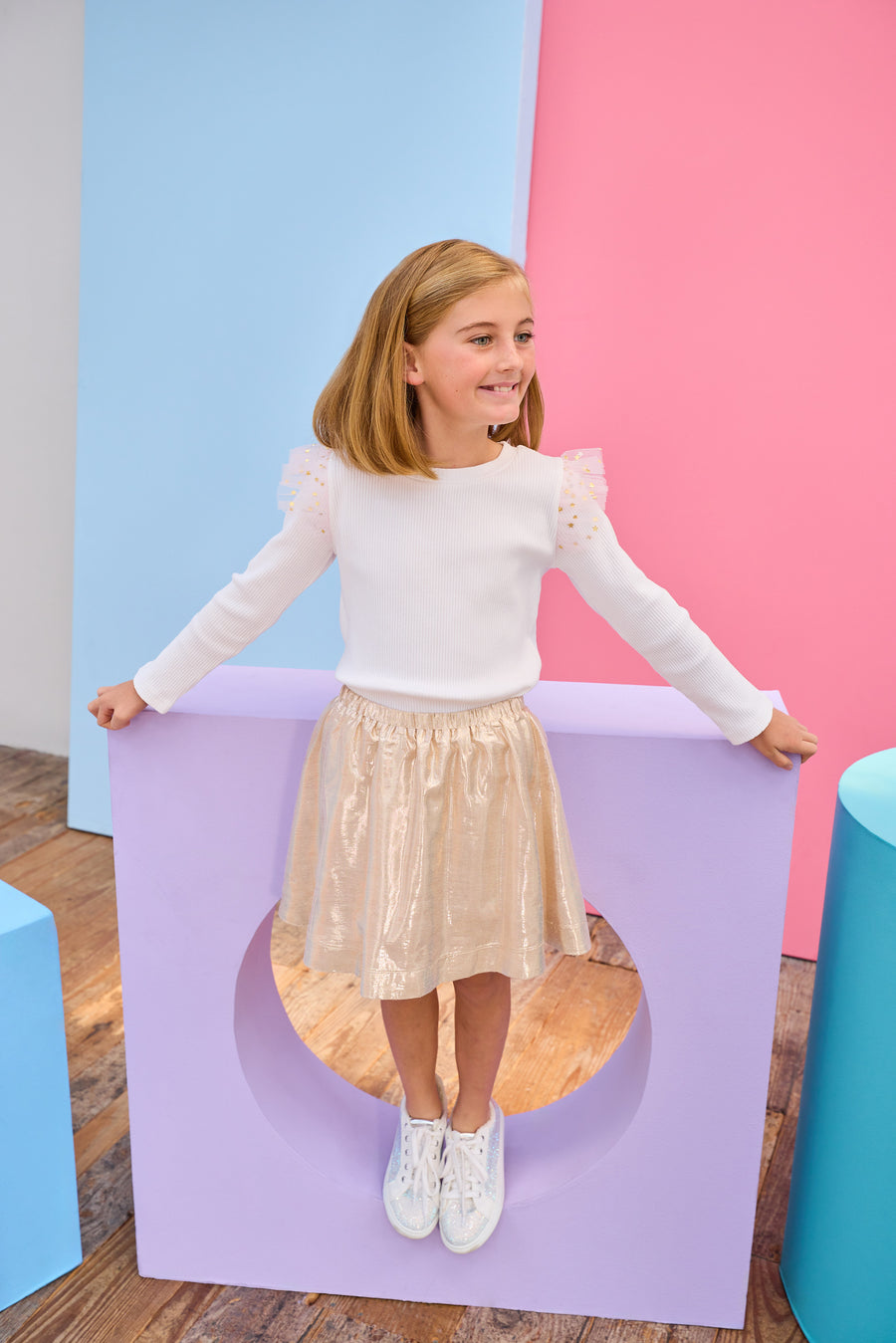 Young girl in a white ribbed long sleeve top gold metallic skirt standing on a purple platform with a colorful background.