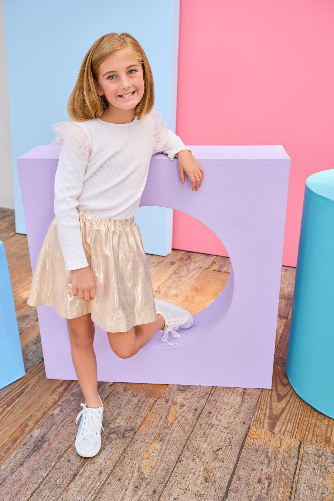 Young girl in a white ribbed long sleeve top gold metallic skirt standing on a purple platform with a colorful background.