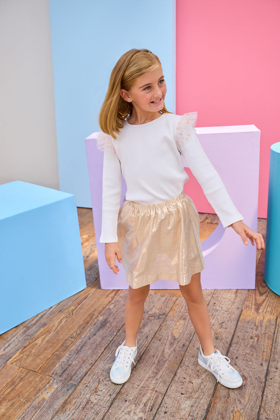 Young girl in a white ribbed long sleeve top gold metallic skirt standing on a purple platform with a colorful background.