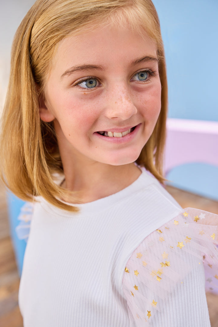 Young girl with blonde hair and blue eyes wearing a white ribbed long sleeve top with pink gold tulle and gold stars at the shoulders 