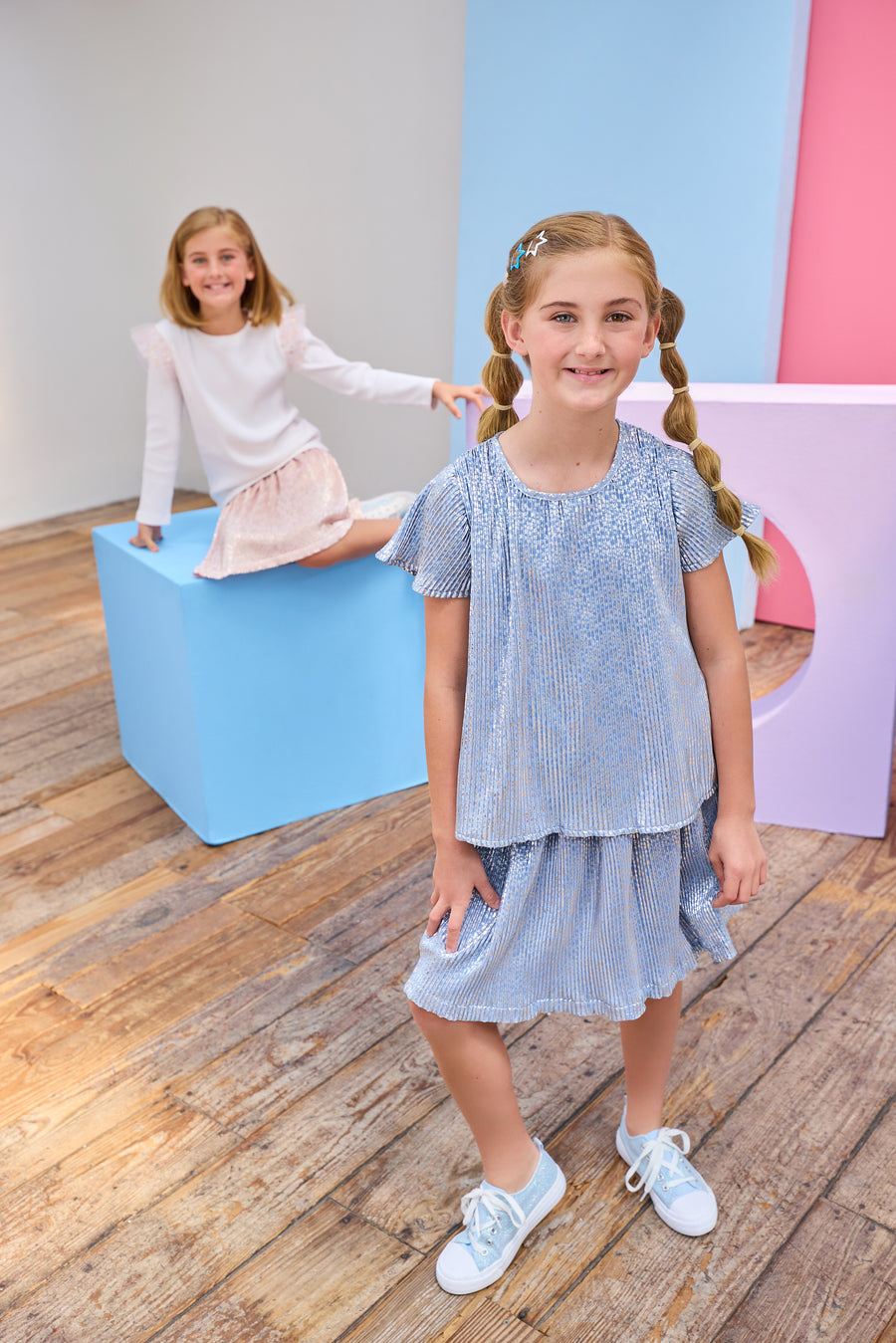 Two young girls in skorts with colorful blocks on a wooden floor.