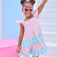 Model can be seen wearing woven pink and blue stripe colorblock pattern. Dress features angel sleeves, smocking across bust, and outside surging detail for an elevated look.