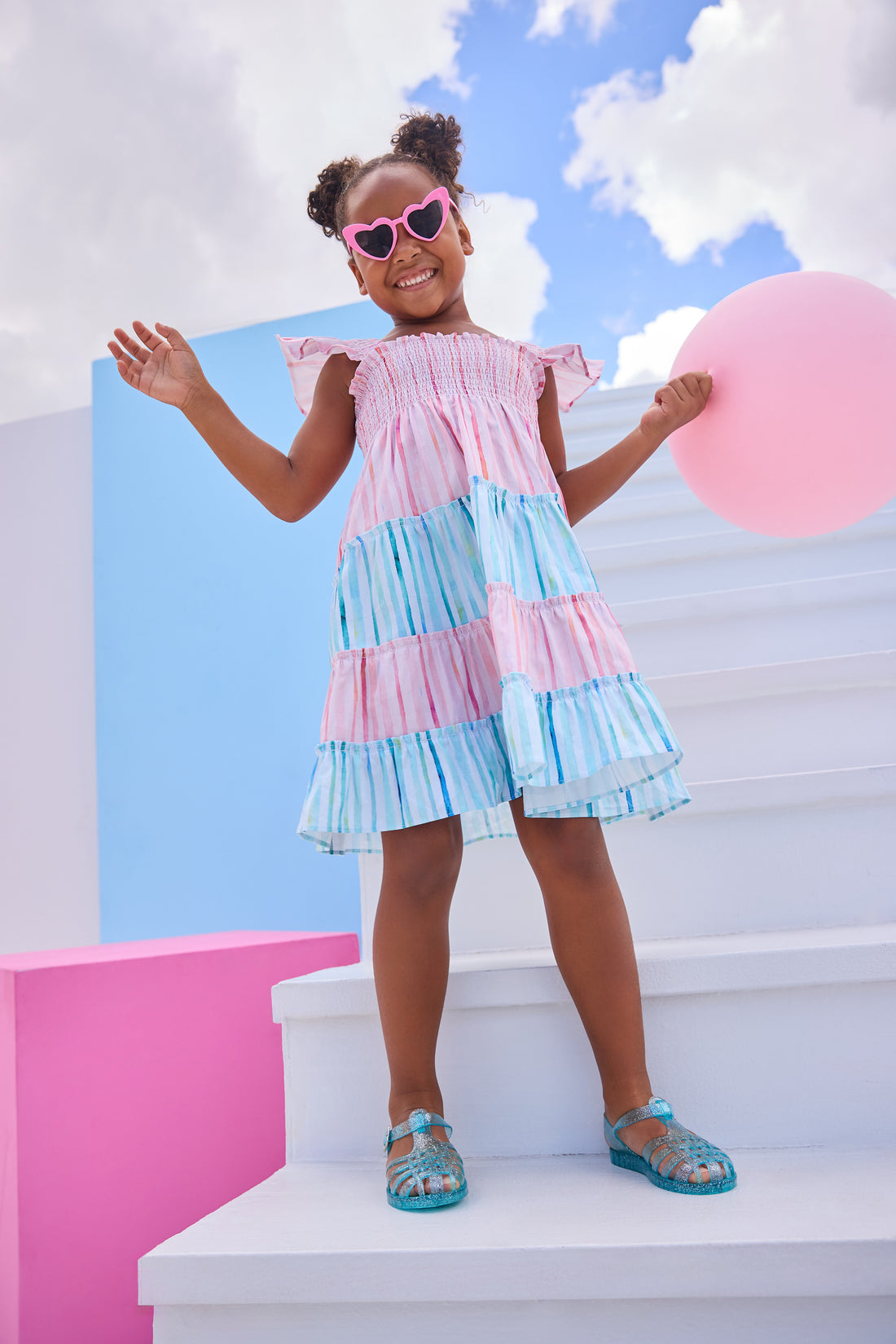 Model can be seen wearing woven pink and blue stripe colorblock pattern. Dress features angel sleeves, smocking across bust, and outside surging detail for an elevated look.