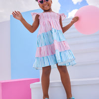 Model can be seen wearing woven pink and blue stripe colorblock pattern. Dress features angel sleeves, smocking across bust, and outside surging detail for an elevated look.