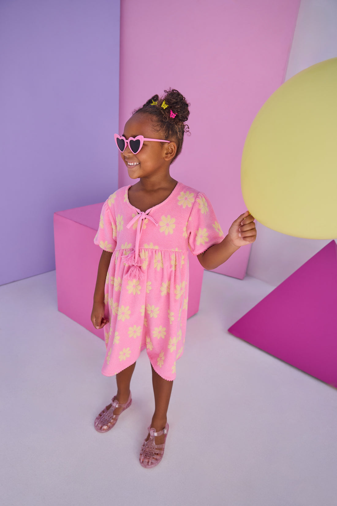 Model can be seen wearing a knit terry material dress in bubblegum pink with yellow flowers. Dress features v-neckline with tassel ties to elevate the look. 