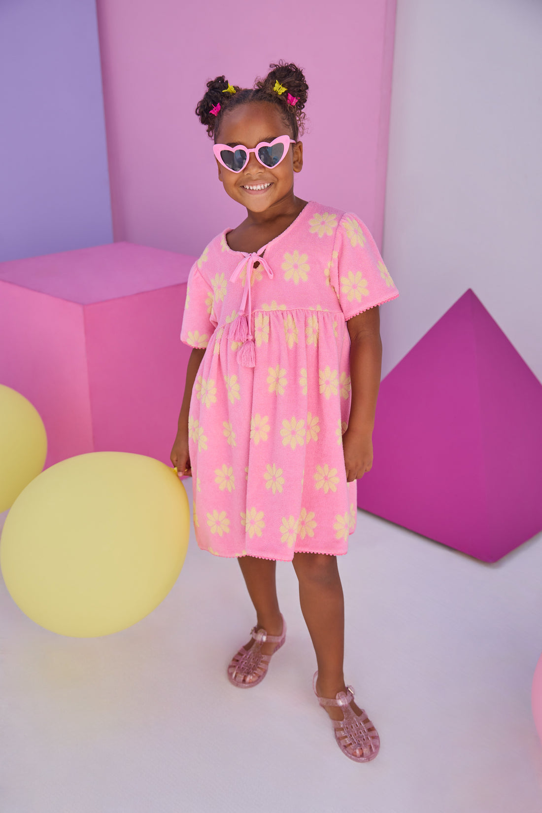 Model can be seen wearing a knit terry material dress in bubblegum pink with yellow flowers. Dress features v-neckline with tassel ties to elevate the look. 