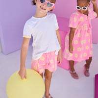 Model can be seen wearing knit terry shorts in a bubblegum pink/yellow floral print. Shorts feature tassel tie detail along with pom pom details along front and side of shorts. Shorts pair back perfectly with white tee with the same yellow/pink colored ric rac detail on the cuffs of sleeves. 