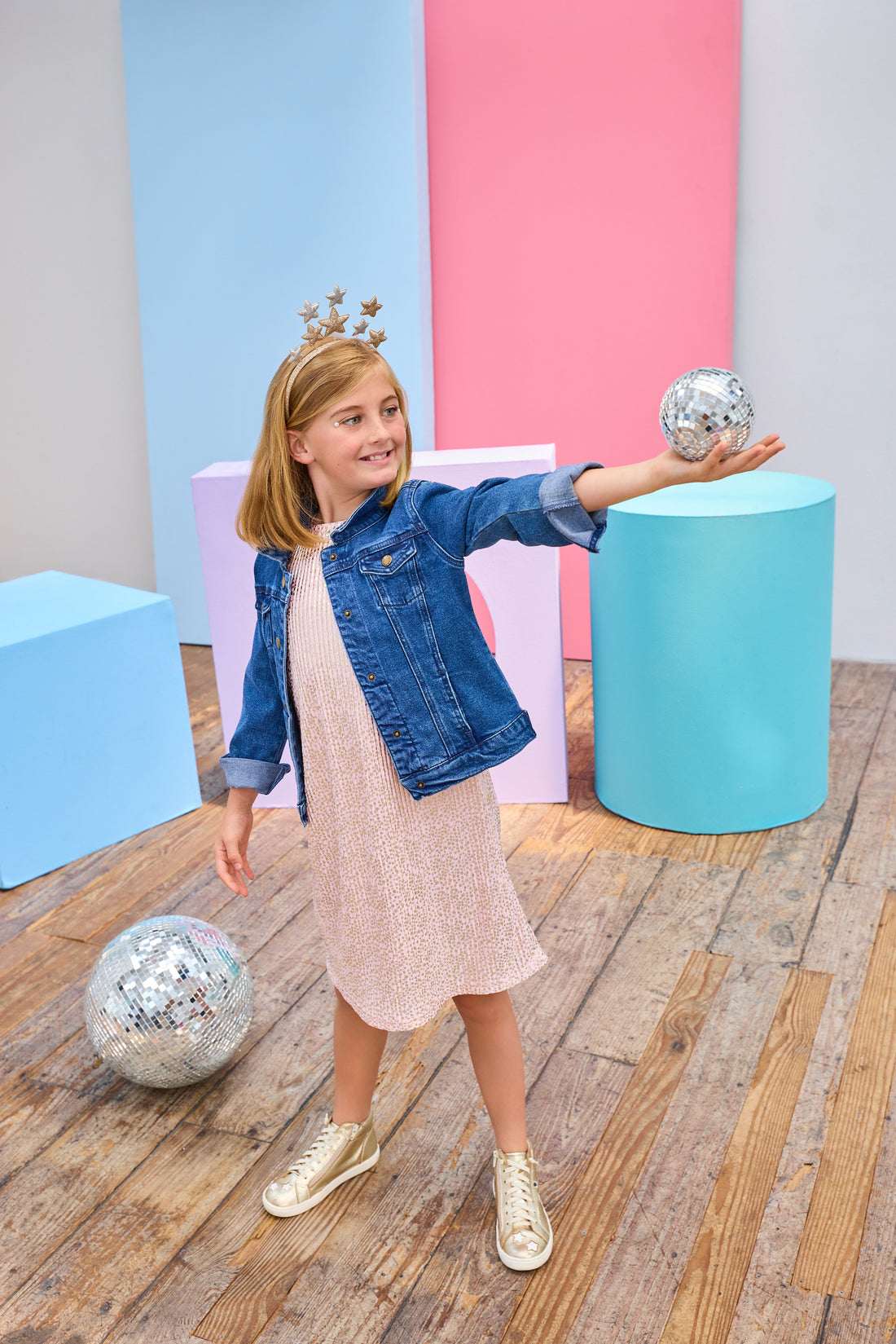 Young girl in a denim jacket and a pink crinkle dress with gold stars holding a disco ball in a colorful room.