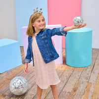 Young girl in a denim jacket and a pink crinkle dress with gold stars holding a disco ball in a colorful room.