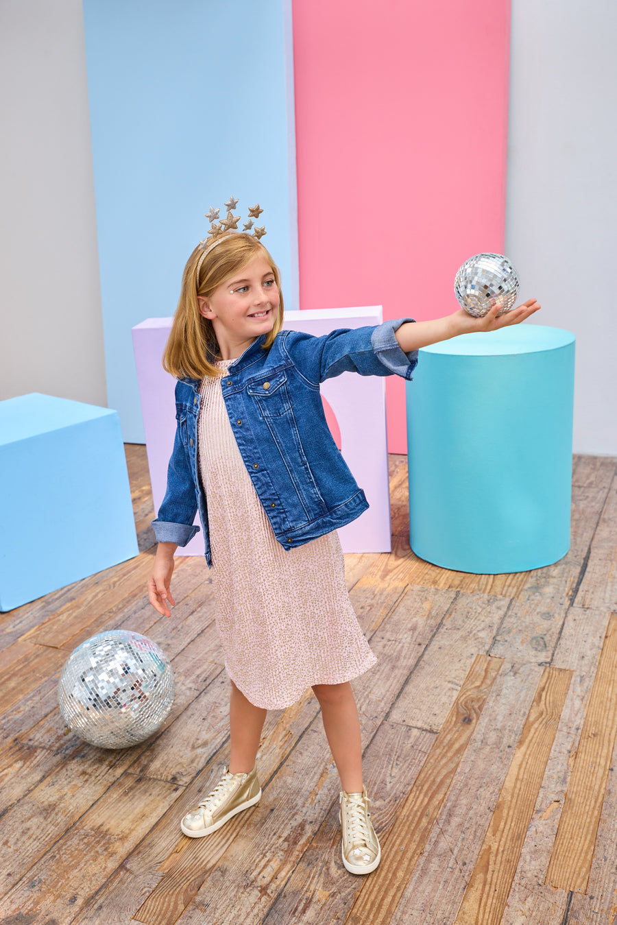 Young girl in a denim jacket and a pink crinkle dress with gold stars holding a disco ball in a colorful room.