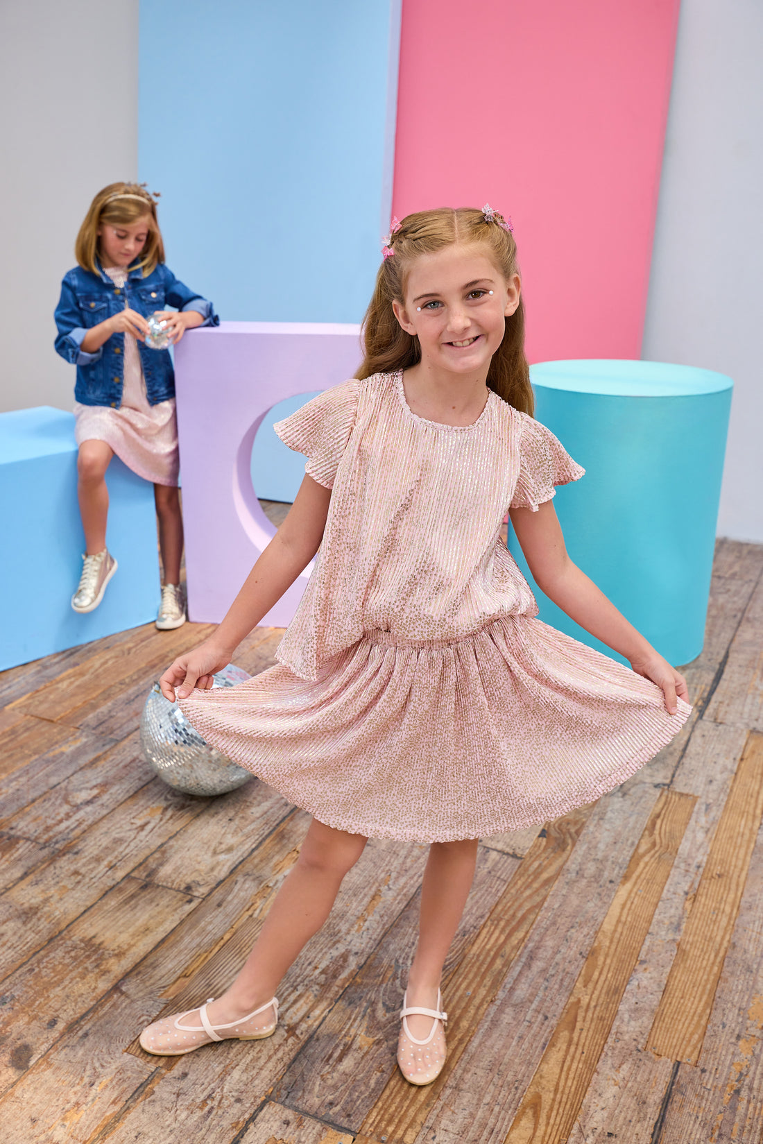 Young girl in a pink crinkle top and skort posing in front of colorful cylindrical objects.