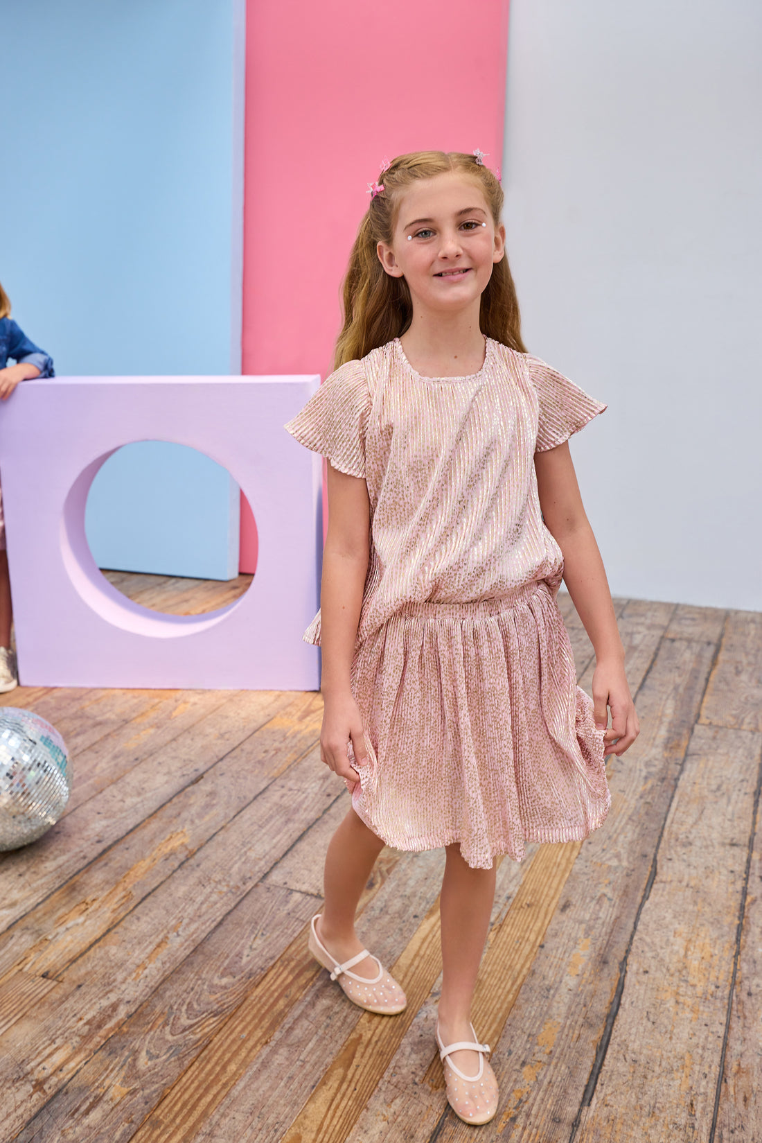 Young girl in a pink crinkle top and skort standing on a wooden floor with colorful geometric shapes in the background.