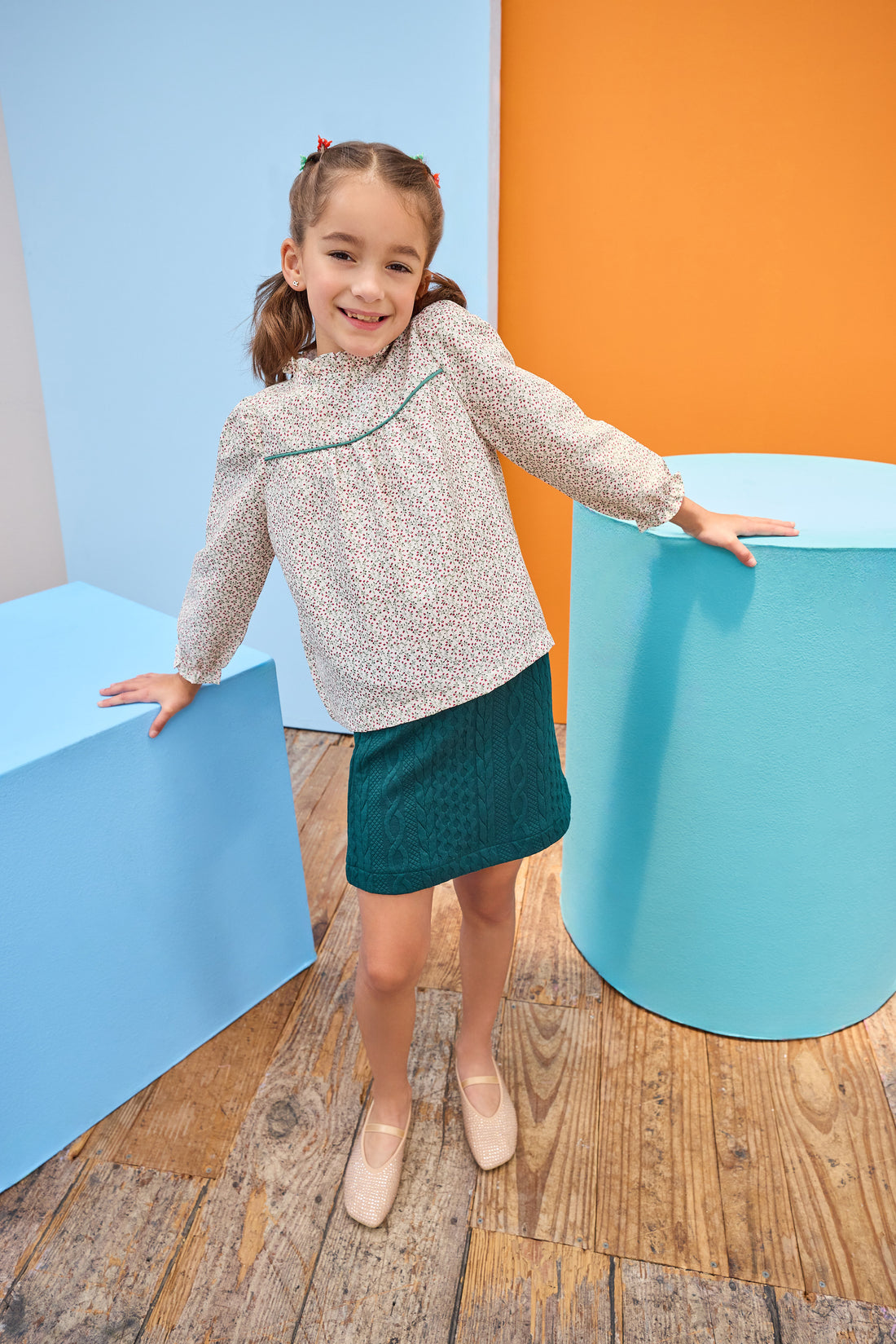 Model can be seen wearing a woven long sleeve top in a red/green floral pattern that features a ruffle high neckline and emerald green trim/pleating detail and button closure along back. Paired back perfectly with a knit green skort to complete the look.