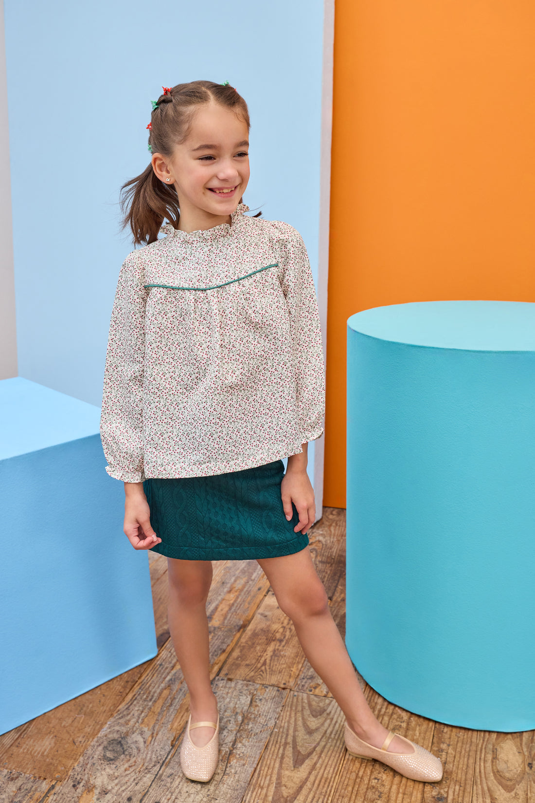 Model can be seen wearing a woven long sleeve top in a red/green floral pattern that features a ruffle high neckline and emerald green trim/pleating detail and button closure along back. Paired back perfectly with a knit green skort to complete the look.