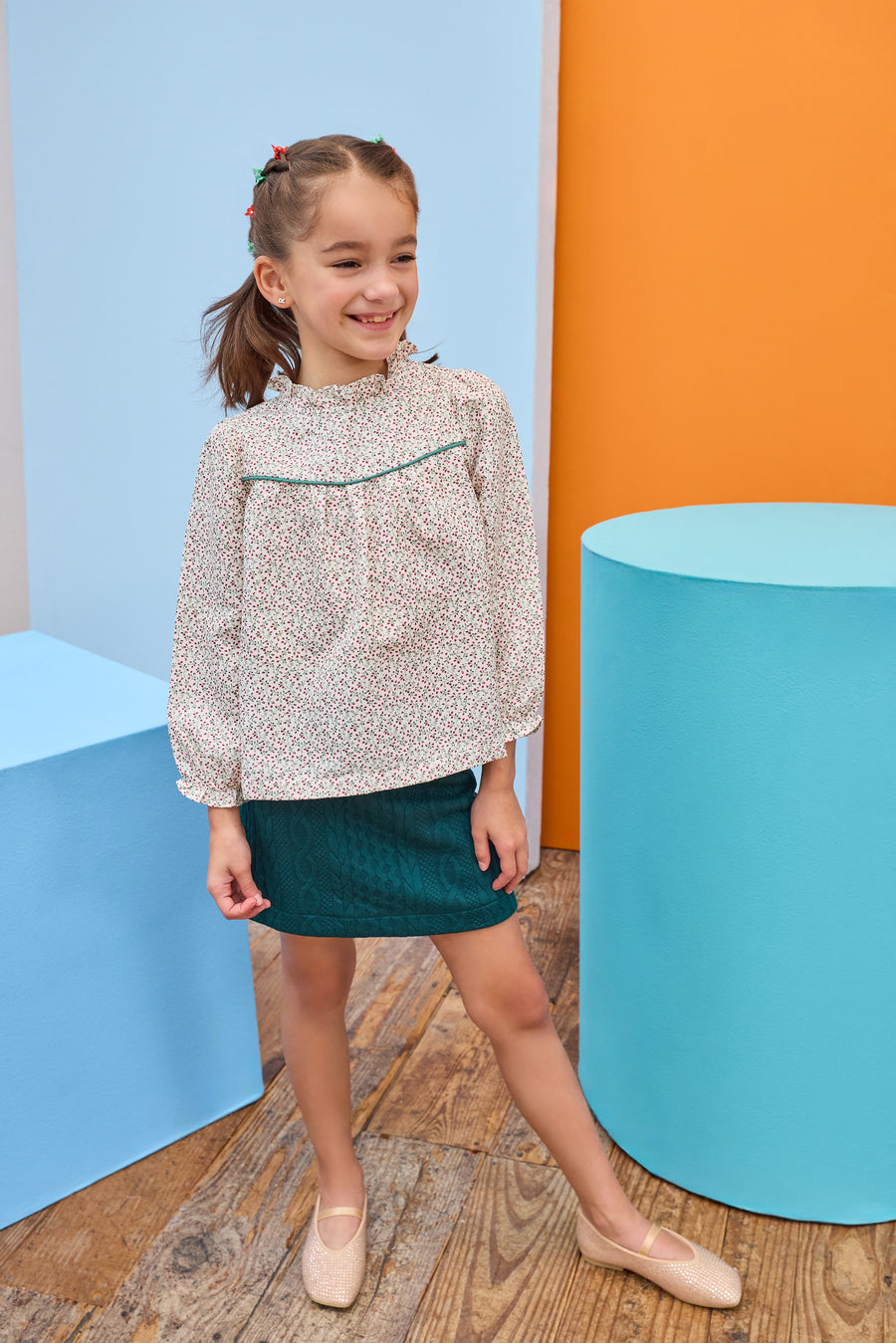 Model can be seen wearing a woven long sleeve top in a red/green floral pattern that features a ruffle high neckline and emerald green trim/pleating detail and button closure along back. Paired back perfectly with a knit green skort to complete the look.