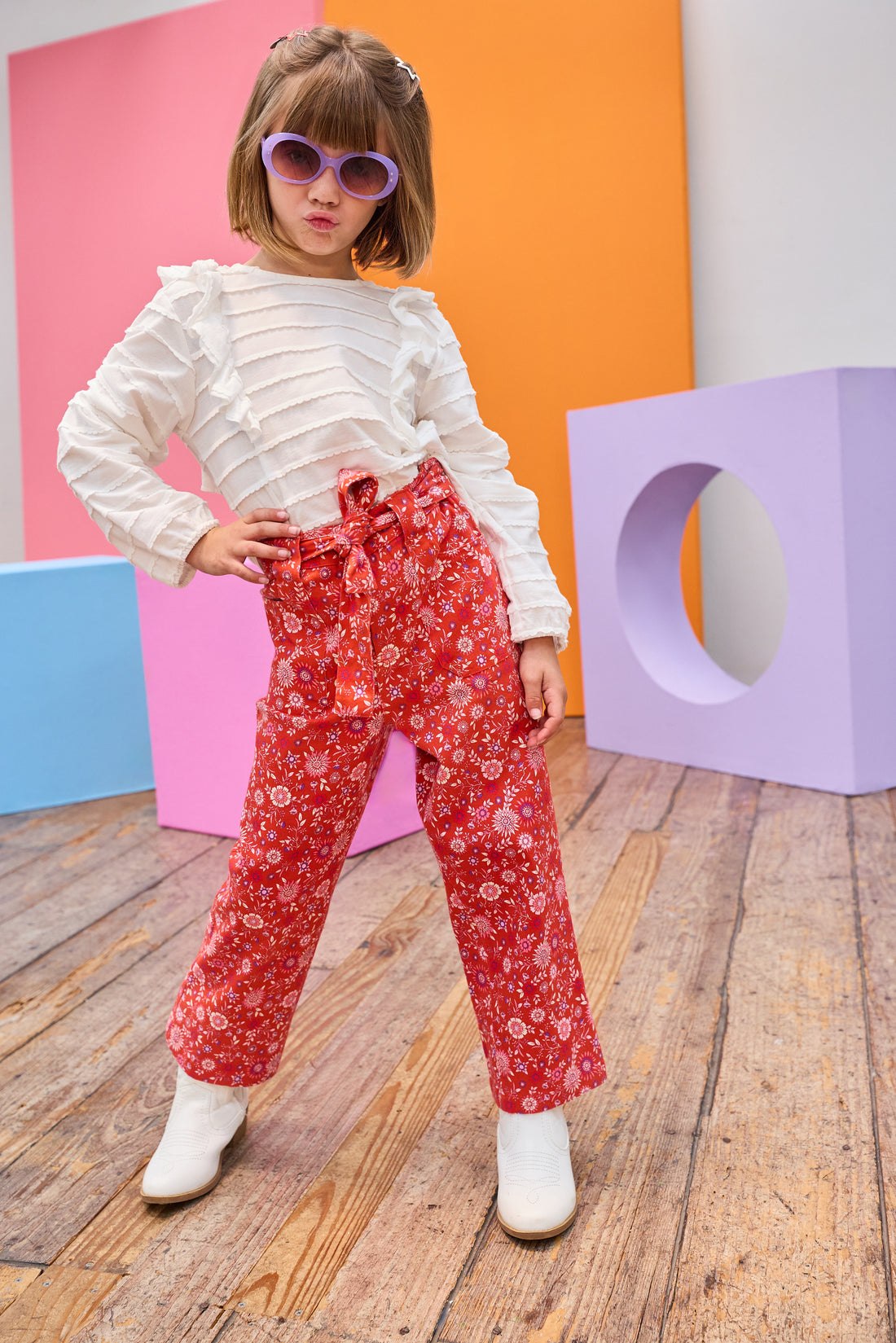 Tween model can be seen wearing an elevated white long sleeve shirt featuring ruffle detail and white scallop detail across the entirety of the shirt paired back with a red and purple floral print pant. Pants feature a belt and zipper/button clasp for a secure fit. 