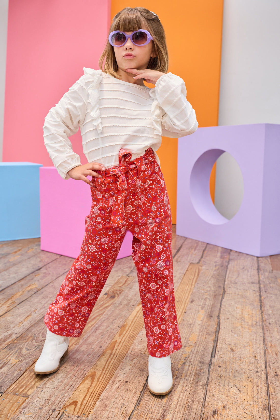 Tween model can be seen wearing an elevated white long sleeve shirt featuring ruffle detail and white scallop detail across the entirety of the shirt paired back with a red and purple floral print pant. Pants feature a belt and zipper/button clasp for a secure fit. 