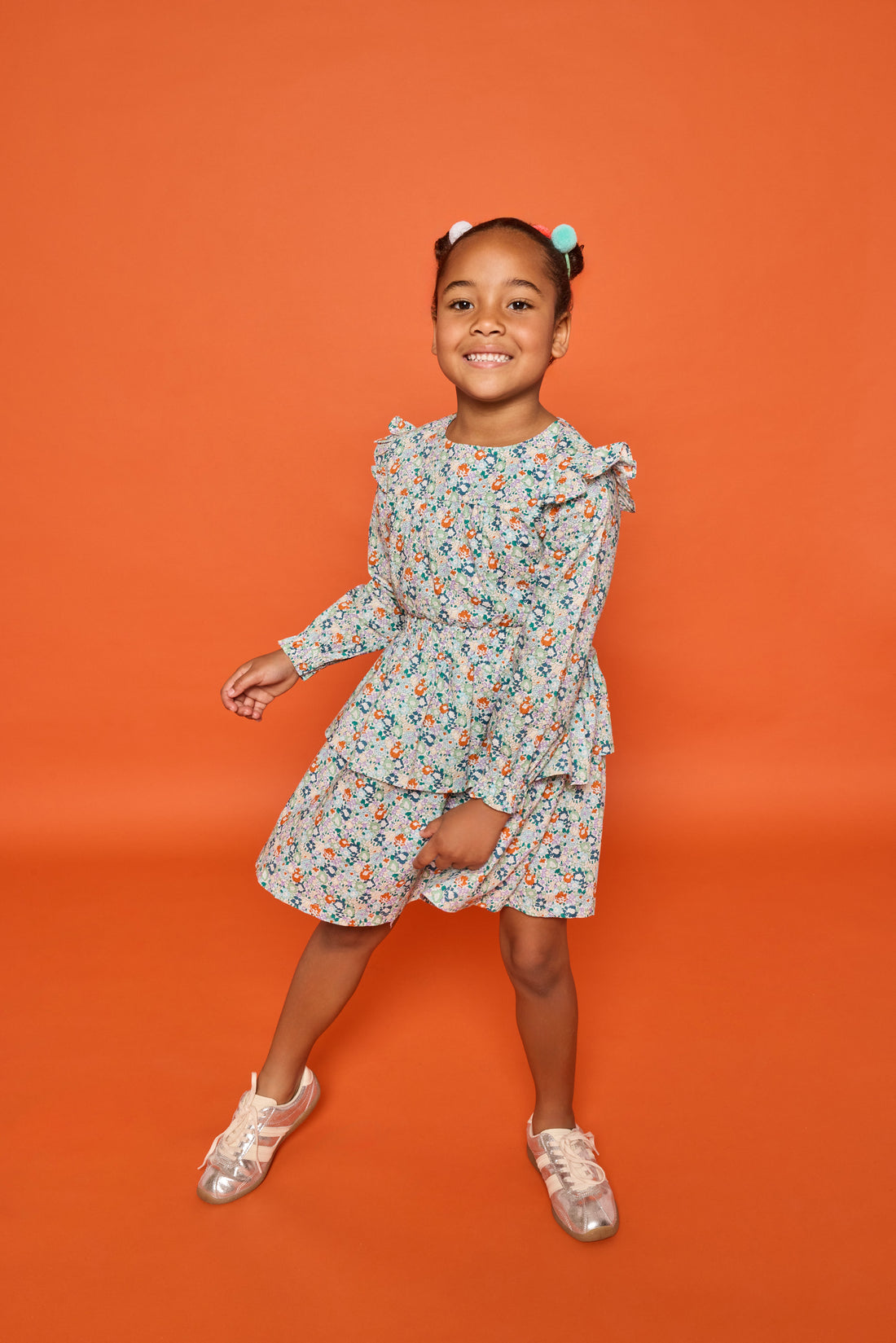 Model can be seen wearing a long sleeve woven dress in an orange/teal/and green floral print. Dress features tiers, elastic ruched waistband, and angel sleeves at the top of the long sleeves for an elevated look. 