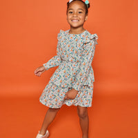 Model can be seen wearing a long sleeve woven dress in an orange/teal/and green floral print. Dress features tiers, elastic ruched waistband, and angel sleeves at the top of the long sleeves for an elevated look. 
