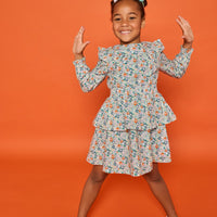 Model can be seen wearing a long sleeve woven dress in an orange/teal/and green floral print. Dress features tiers, elastic ruched waistband, and angel sleeves at the top of the long sleeves for an elevated look. 