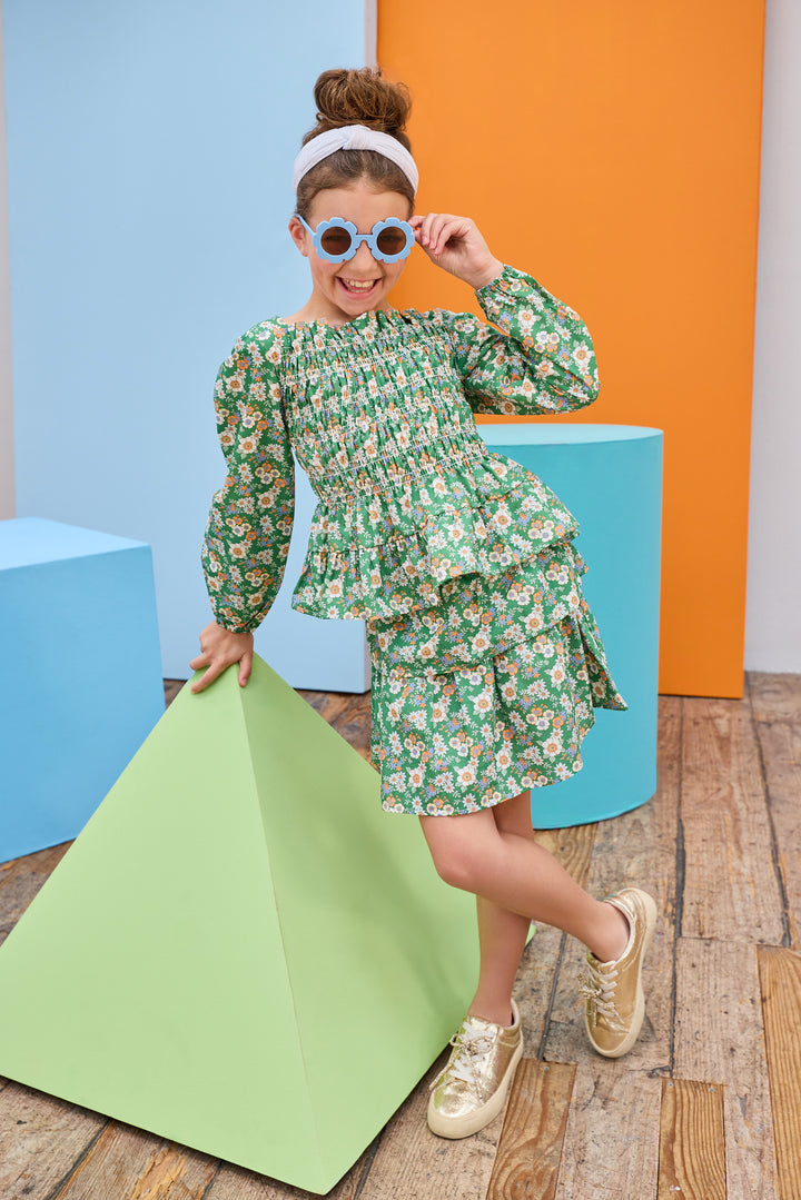 Model can be seen wearing a long sleeve woven top in a green/orange/blue floral print,  that features smocking detail throughout body of shirt for a form fitting but stretchy fit. Top also pairs back perfectly with the matching two tiered skort in the same print to complete the look. 