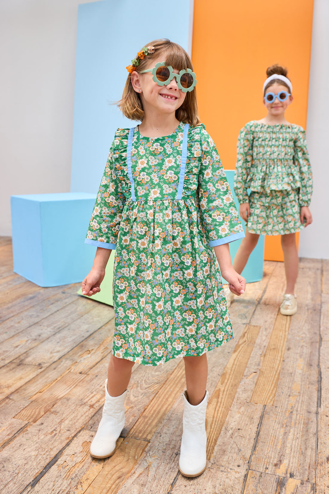 Model can be seen wearing a long sleeve woven dress in a beautiful green/blue/orange floral print. Dress features button closure along back,  ruffle detail on outside of bust, and blue ribbon trim along ruffle line/sleeve trim to elevate the look. 