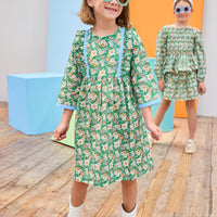 Model can be seen wearing a long sleeve woven dress in a beautiful green/blue/orange floral print. Dress features button closure along back,  ruffle detail on outside of bust, and blue ribbon trim along ruffle line/sleeve trim to elevate the look. 
