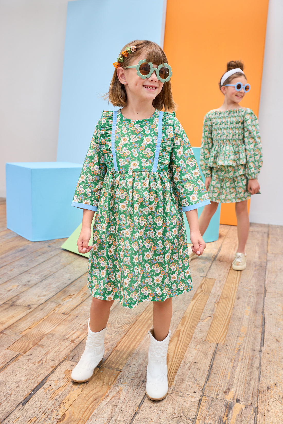 Model can be seen wearing a long sleeve woven dress in a beautiful green/blue/orange floral print. Dress features button closure along back,  ruffle detail on outside of bust, and blue ribbon trim along ruffle line/sleeve trim to elevate the look. 