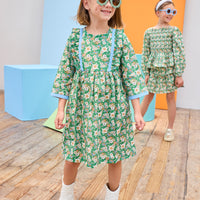 Model can be seen wearing a long sleeve woven dress in a beautiful green/blue/orange floral print. Dress features button closure along back,  ruffle detail on outside of bust, and blue ribbon trim along ruffle line/sleeve trim to elevate the look. 