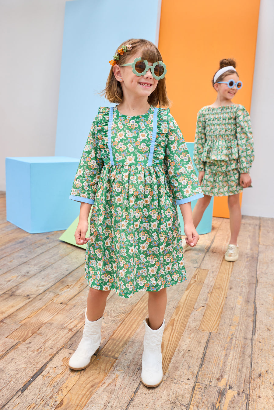 Model can be seen wearing a long sleeve woven dress in a beautiful green/blue/orange floral print. Dress features button closure along back,  ruffle detail on outside of bust, and blue ribbon trim along ruffle line/sleeve trim to elevate the look. 