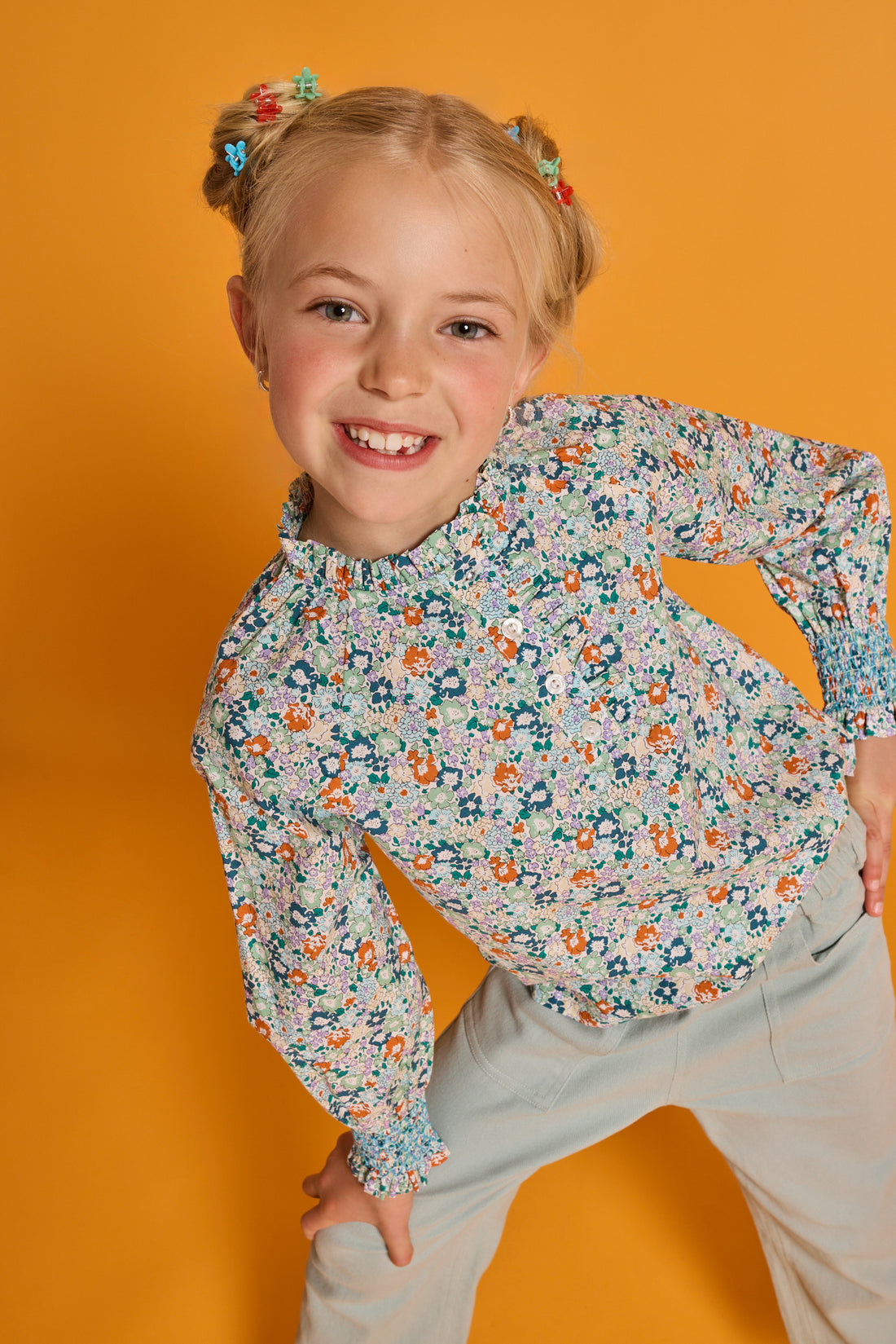 Model can be seen wearing an orange/green/teal floral print woven shirt that features buttons down the front with ruffled detail along neckline and smocking detail at sleeves for an elevated look. Paired back perfectly with our light green pants with elastic waistband and pockets in the front. 