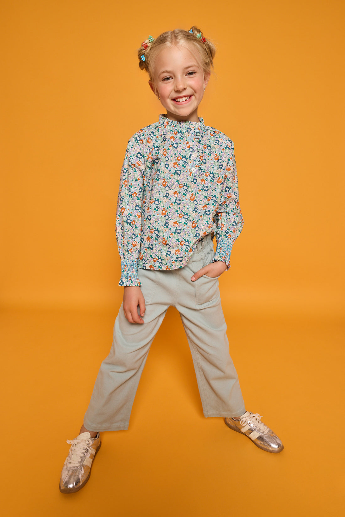 Model can be seen wearing an orange/green/teal floral print woven shirt that features buttons down the front with ruffled detail along neckline and smocking detail at sleeves for an elevated look. Paired back perfectly with our light green pants with elastic waistband and pockets in the front. 