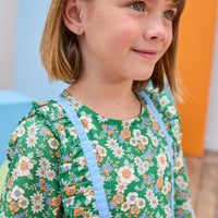 Model can be seen wearing a long sleeve woven dress in a beautiful green/blue/orange floral print. Dress features button closure along back,  ruffle detail on outside of bust, and blue ribbon trim along ruffle line/sleeve trim to elevate the look. 
