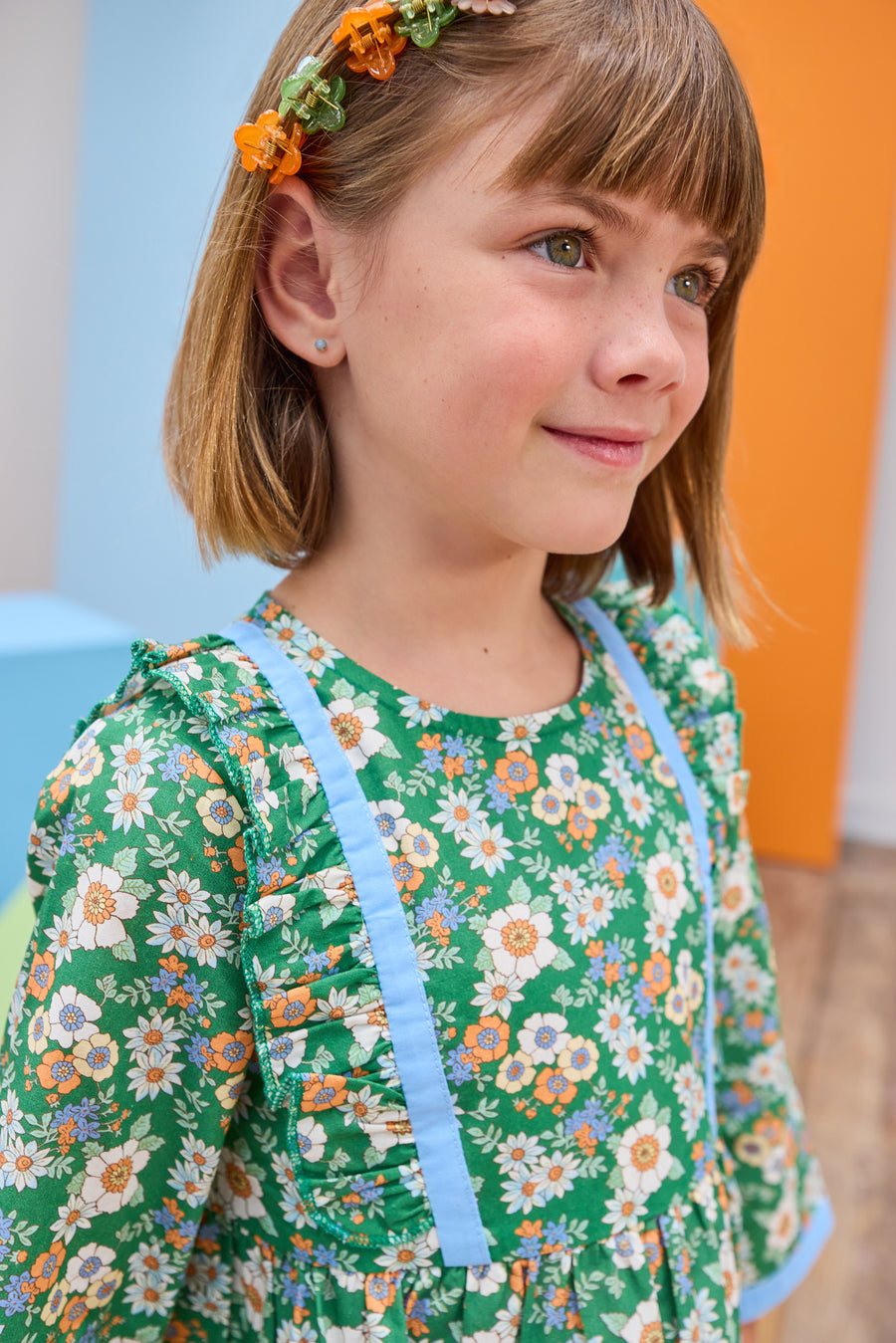 Model can be seen wearing a long sleeve woven dress in a beautiful green/blue/orange floral print. Dress features button closure along back,  ruffle detail on outside of bust, and blue ribbon trim along ruffle line/sleeve trim to elevate the look. 