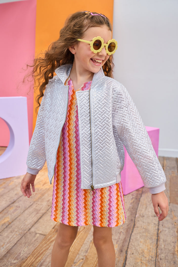 Young girl wearing a colorful dress and a silver quilted jacket with yellow sunglasses, standing on a wooden floor with a colorful abstract background.