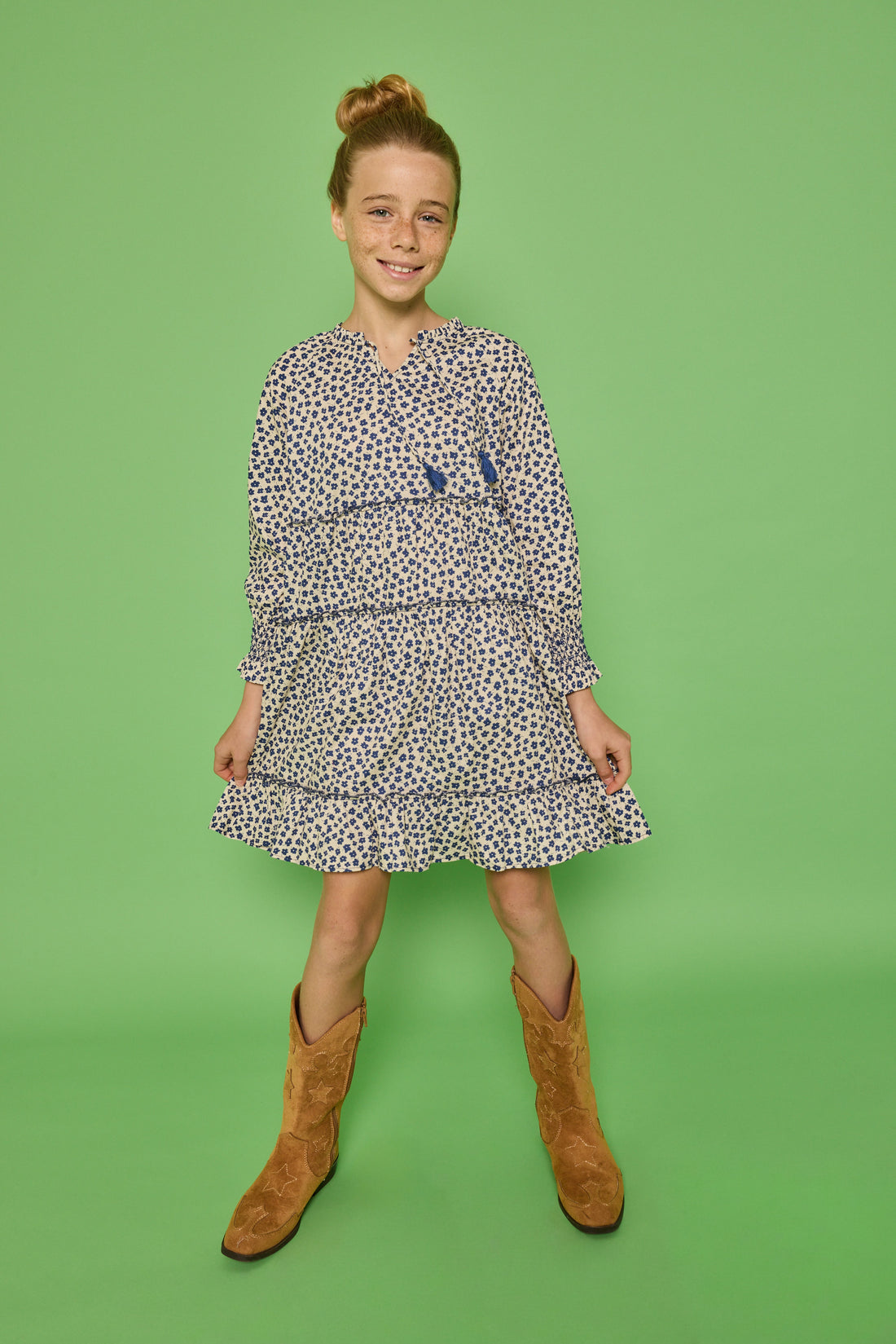 Model can be seen wearing a long sleeve woven dress in a blue/cream floral print. Dress features tier detail along body, elastic cuffs at sleeves, and v-neckline with tassel detail to complete the look. 