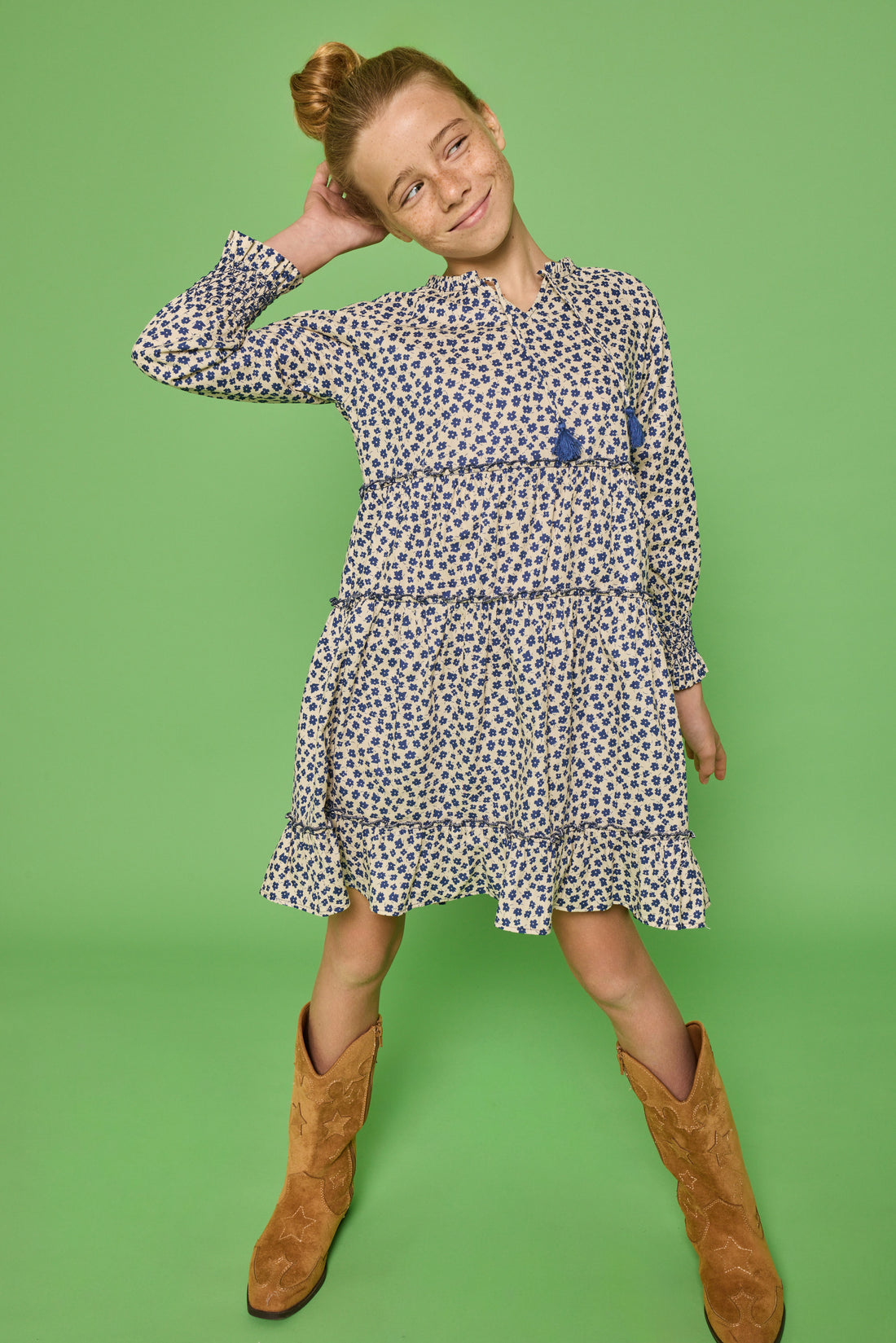 Model can be seen wearing a long sleeve woven dress in a blue/cream floral print. Dress features tier detail along body, elastic cuffs at sleeves, and v-neckline with tassel detail to complete the look. 