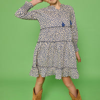 Model can be seen wearing a long sleeve woven dress in a blue/cream floral print. Dress features tier detail along body, elastic cuffs at sleeves, and v-neckline with tassel detail to complete the look. 