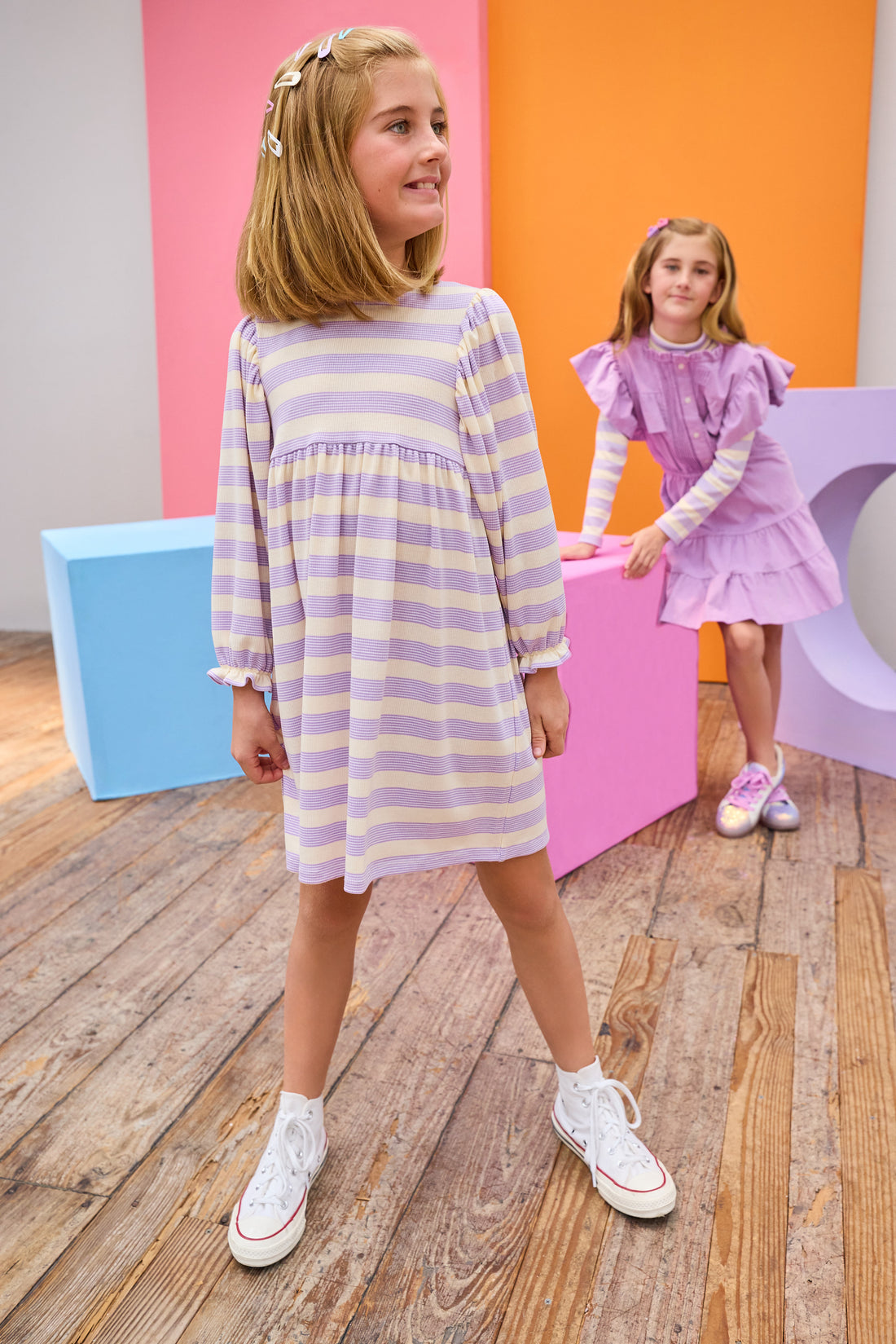Two young girls in purple dresses standing in a room with a wooden floor and colorful walls.