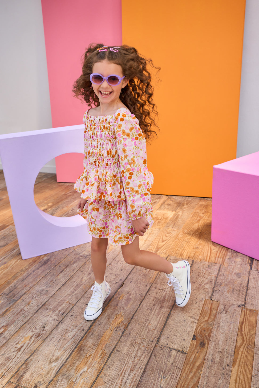 Child in a floral top and skirt wearing sunglasses posing in front of colorful geometric shapes.