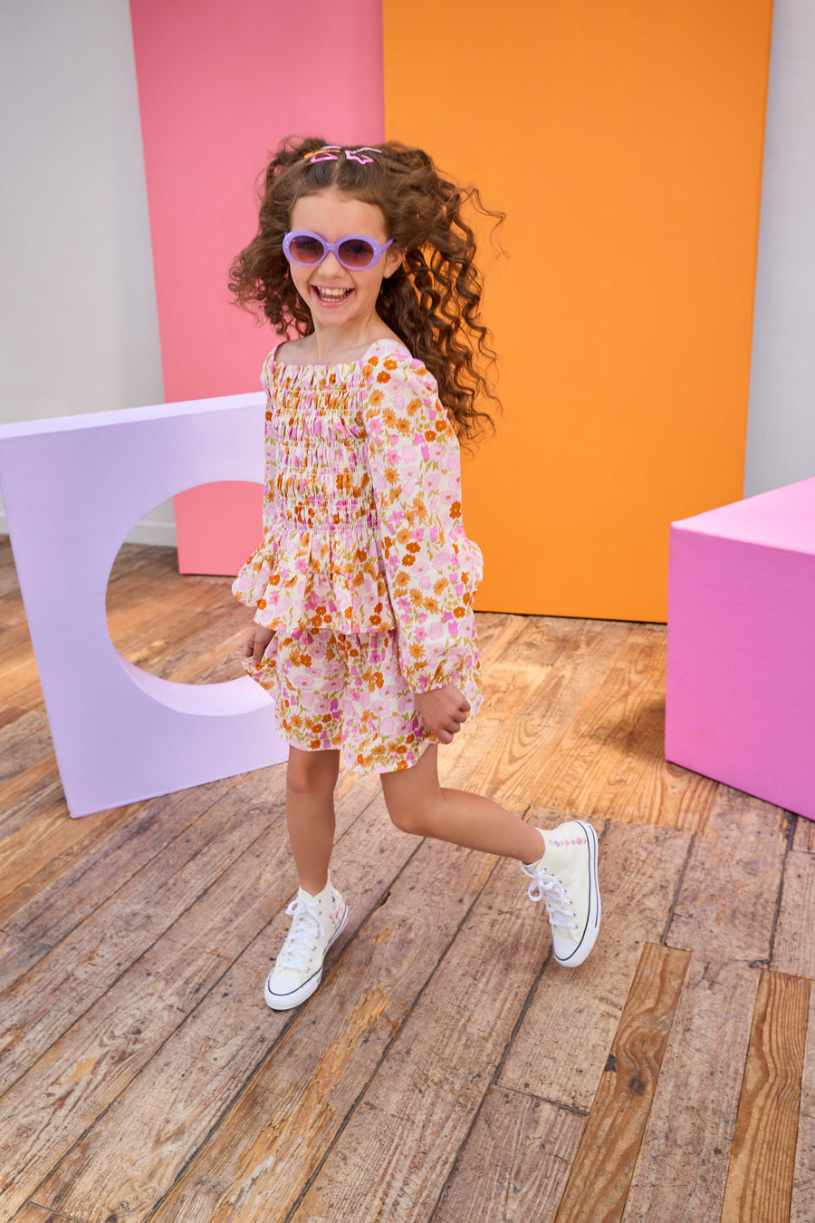 Child in a floral top and skirt wearing sunglasses posing in front of colorful geometric shapes.