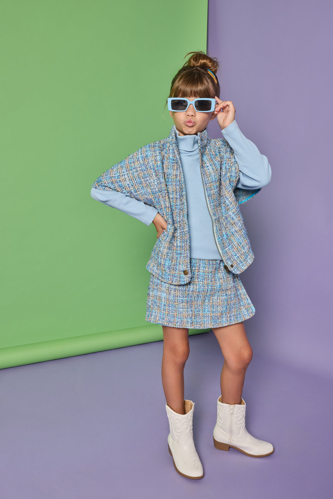 Model can be seen wearing a tweed cape in a blue tweed pattern. Cape pairs back with the matching tweed skort and ribbed turtleneck underneath to complete the look.